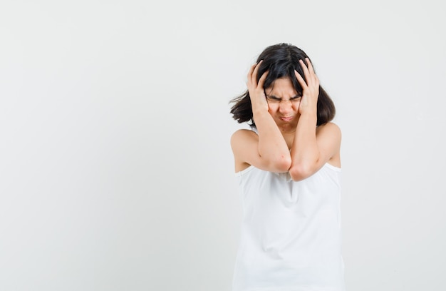 Little girl clasping head in hands in white blouse and looking irritated , front view.