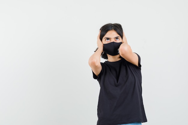 Little girl clasping head with hands in black t-shirt, mask and looking scared. front view.