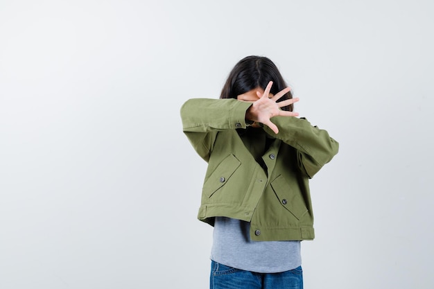 Маленькая девочка в пальто, футболке, джинсах, закрывающих глаза рукой, показывая ладонь и выглядя испуганной, вид спереди.