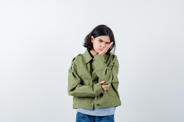 Маленькая девочка в пальто, футболке, джинсах, прислонившись щекой к руке и задумчиво, вид спереди.