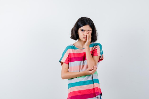 Little girl covering eye with hand while holding hand on elbow, looking away in t-shirt and looking sleepy , front view.