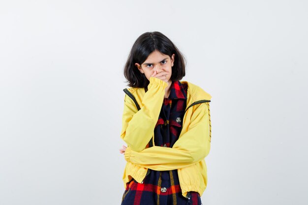 Little girl covering mouth with hand in checked shirt, jacket and looking amazed , front view.