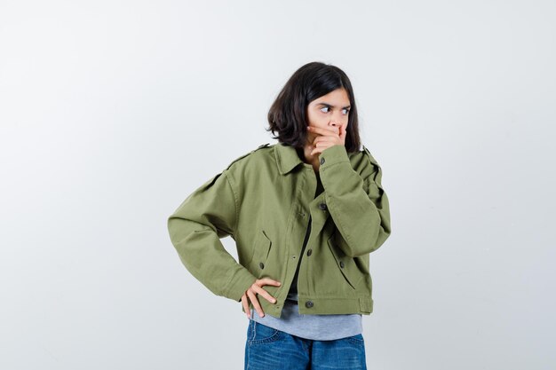 Little girl covering mouth with hand in coat, t-shirt, jeans and looking amazed. front view.