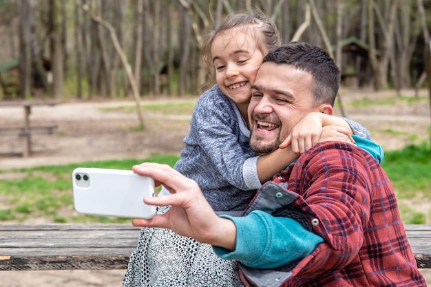 Маленькая девочка и папа фотографируются на переднем плане мобильного телефона в парке ранней весной.
