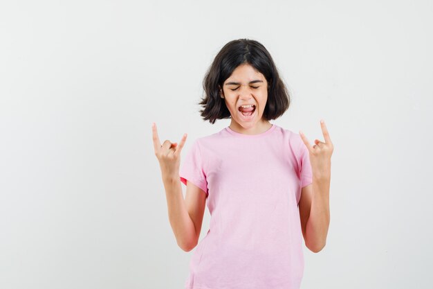 Маленькая девочка делает рок-символ, крича в розовой футболке и выглядит энергичным, вид спереди.