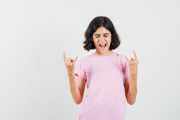 Little girl doing rock symbol while screaming in pink t-shirt and looking energetic , front view.