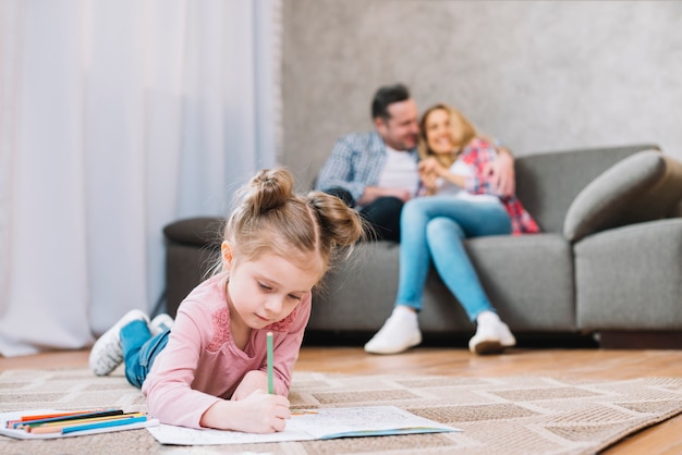 Little girl drawing on book lying floor while her loving parents sitting on sofa