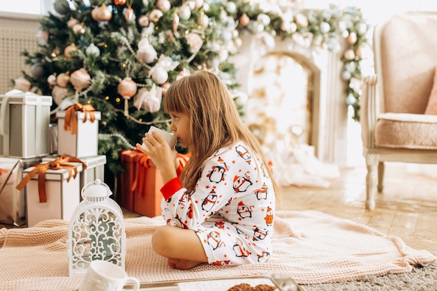 Маленькая девочка в пижаме сидит на ковре и пьет какао с зефиром возле новогодней елки в уютной комнате.