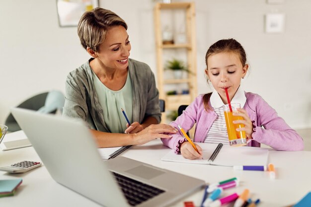 Little girl drinking juice while doing homework with her mother