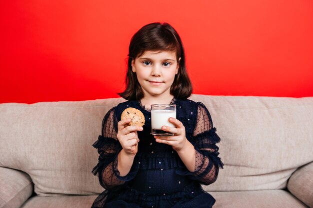 赤い壁に牛乳を飲む少女。クッキーを食べる子供の屋内ショット。