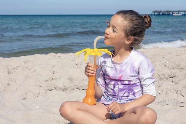 Little girl drinks juice sitting on the sand near the sea