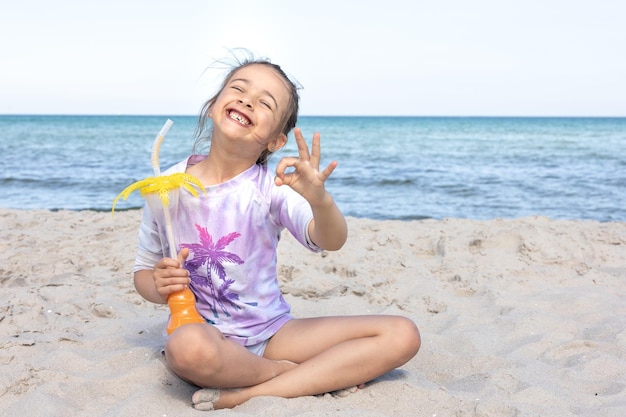 Little girl drinks juice sitting on the sand near the sea