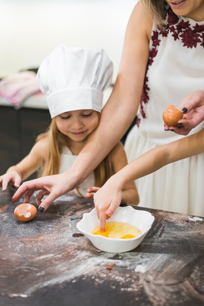 어머니가 지저분한 부엌 카운터에 음식을 준비하는 동안 계란에 어린 소녀 dunking 손가락