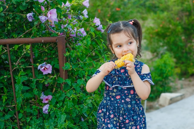 Little girl eating fresh corn while standing in spring dress in garden , front view.