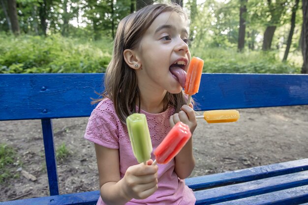 A little girl eats a colorful ice cream made from frozen popsicles on a bench on a hot summer day.
