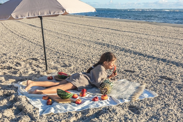 少女はビーチで毛布の上に横たわって果物を食べる