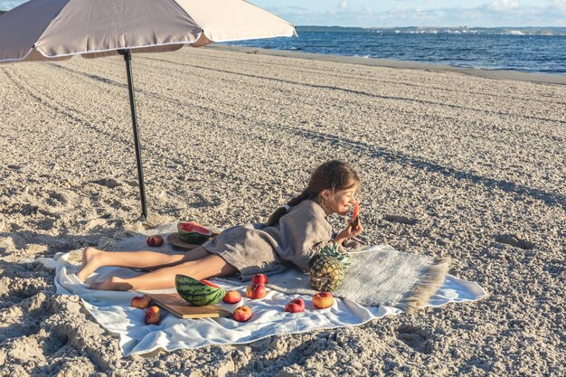 少女はビーチで毛布の上に横たわって果物を食べる