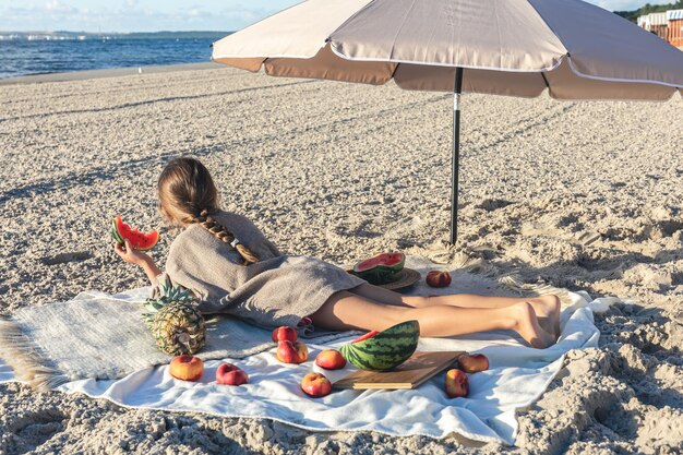 Little girl eats fruit lying on a blanket on the beach