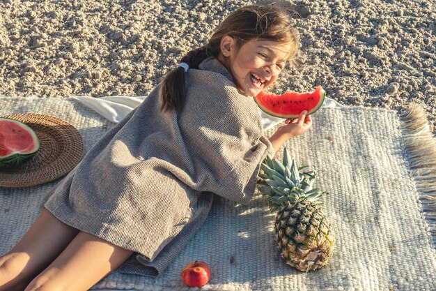 少女はビーチで毛布の上に横たわって果物を食べる