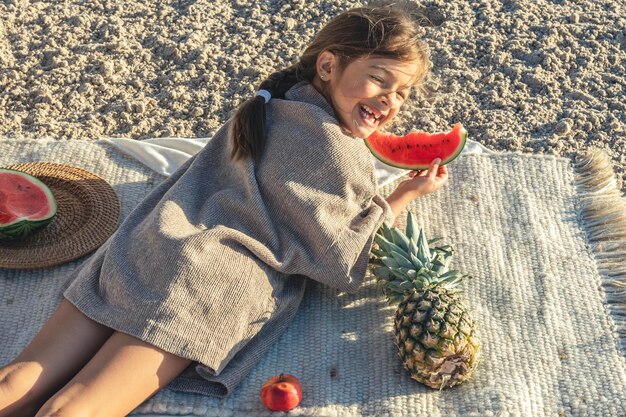 Little girl eats fruit lying on a blanket on the beach