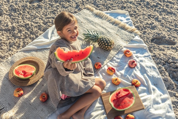 少女はビーチで毛布の上に横たわって果物を食べる