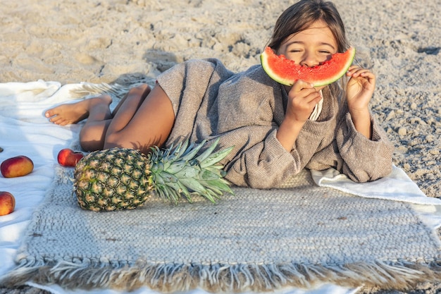 어린 소녀는 해변에서 담요에 누워 과일을 먹는다