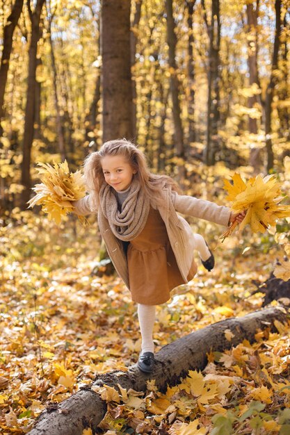 Little girl in fashion clothes standing in autumn forest. Girl holding a yellow leaves. Girl wearing brown dress and a coat.