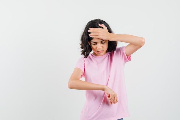 Little girl gesturing like looking at watch on wrist in pink t-shirt and looking anxious , front view.