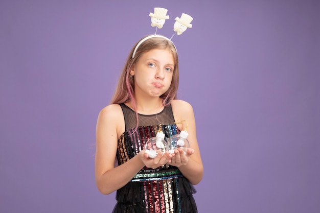 Маленькая девочка в блестящем вечернем платье и забавной повязке на голову держит елочные шары, недовольно глядя в камеру, стоя на фиолетовом фоне