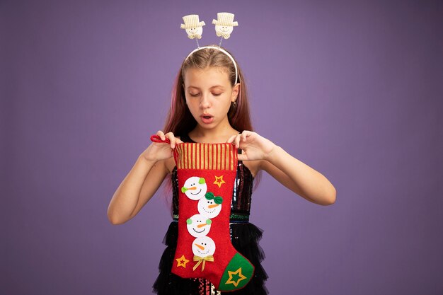 반짝이 파티 드레스와 보라색 배경 위에 서있는 흥미 진진한 내부 찾고 크리스마스 스타킹을 들고 재미있는 머리띠에 어린 소녀