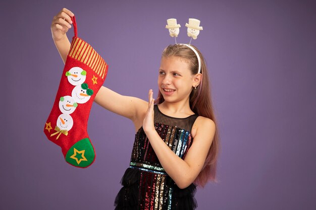 キラキラパーティードレスとそれを見てクリスマスの靴下を保持している面白いヘッドバンドの少女は、紫色の背景の上に立って手を握って不満を持っています