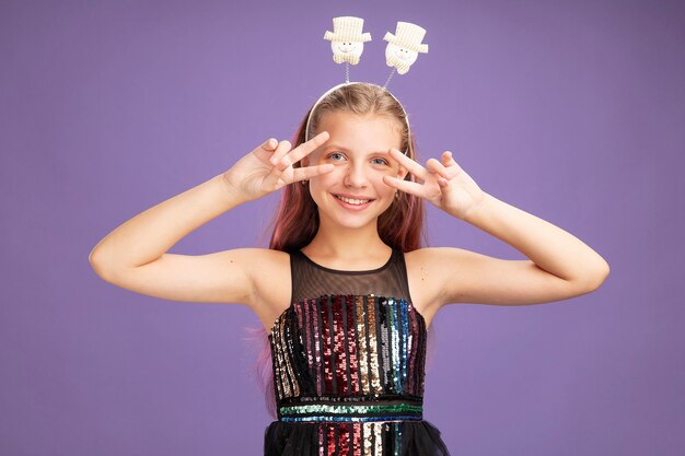 Little girl in glitter party dress and funny headband looking at camera showing v-sign near ger eyes smiling cheerfully standing over purple background