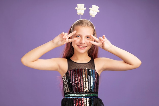 Little girl in glitter party dress and funny headband looking at camera showing v-sign near ger eyes smiling cheerfully standing over purple background