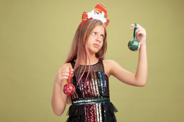반짝이 파티 드레스와 산타 크리스마스 공을 들고 머리띠에 어린 소녀 녹색 배경 위에 서있는 선택을 시도하는 데 의심을 갖는 혼란 찾고