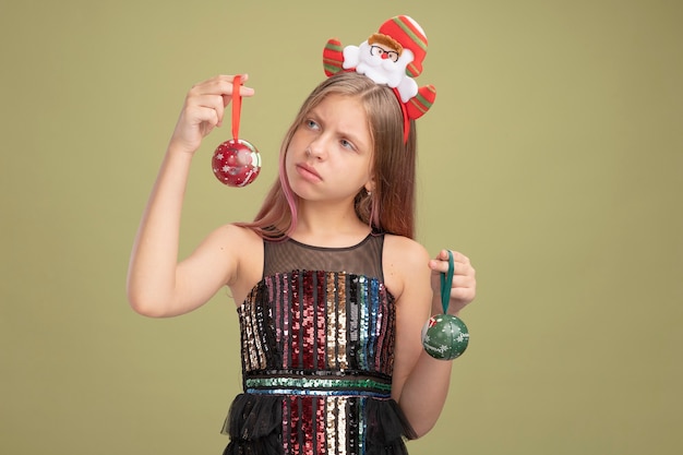 Bambina in abito da festa glitterato e fascia con babbo natale che tiene le palle di natale guardandole confuse avendo dubbi in piedi su sfondo verde