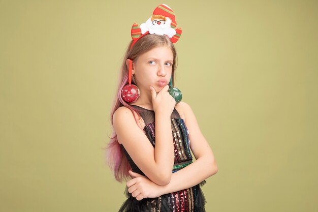 Little girl in glitter party dress and headband with santa with christmas balls on her ears looking aside with hand on chin thinking with serious face standing over green wall