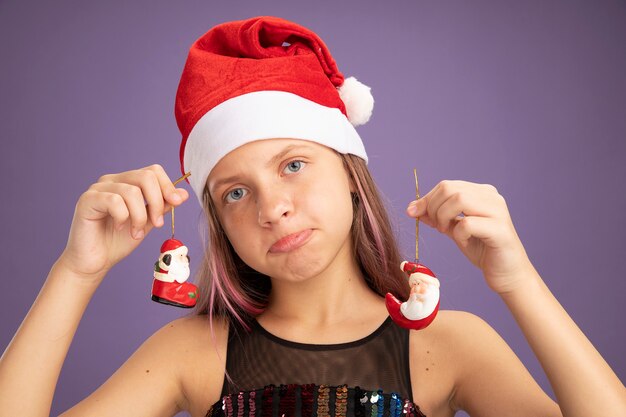 Маленькая девочка в блестящем праздничном платье и шляпе санта-клауса с рождественскими игрушками смотрит в камеру с грустным выражением лица, поджимая губы, стоя на фиолетовом фоне