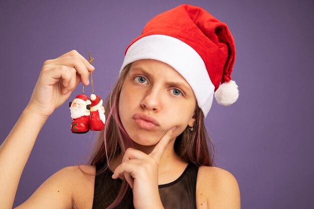 Little girl in glitter party dress and santa hat holding christmas toys looking at camera with serious face standing over purple background