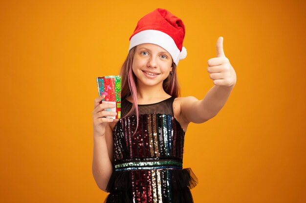 Little girl in glitter party dress and santa hat holding two colorful paper cup looking at camera with smile on face showing thumbs up standing over orange background