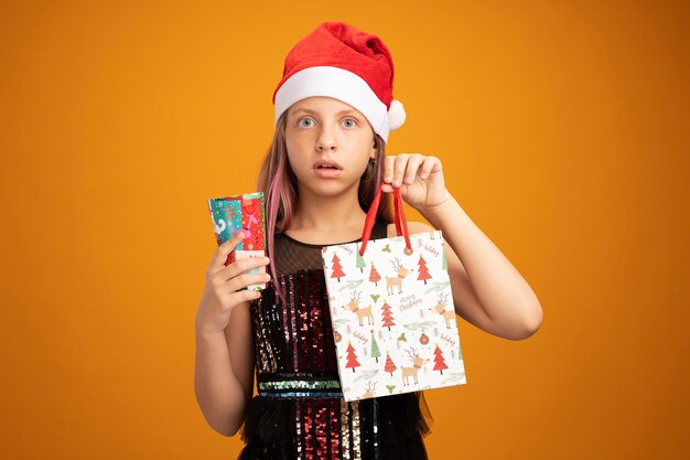 Little girl in glitter party dress and santa hat holding two colorful paper cup and paper bag with gifts looking at camera surprised standing over orange background