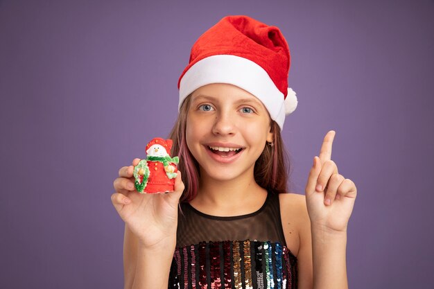 Little girl in glitter party dress and santa hat showing christmas toy looking at camera happy and surprised showing index finger standing over purple background