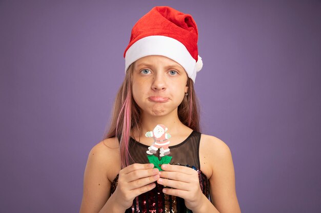 Little girl in glitter party dress and santa hat showing christmas toy looking at camera with sad expression pursing lips standing over purple background