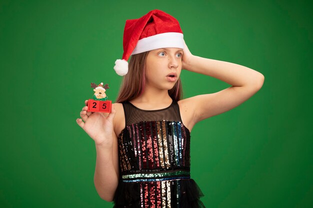 Маленькая девочка в блестящем вечернем платье и шляпе санта-клауса показывает игрушечные кубики с датой двадцать пять, удивленно глядя в сторону, стоя на зеленом фоне