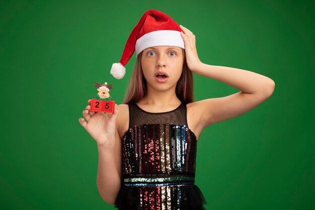 반짝이 파티 드레스와 산타 모자에있는 어린 소녀는 녹색 배경 위에 서있는 그녀의 머리에 손으로 놀란 카메라를보고 25 날짜와 장난감 큐브를 보여주는 산타 모자