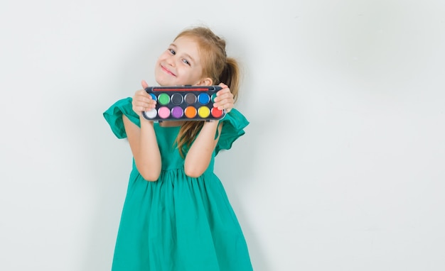 Free photo little girl in green dress holding watercolor paints with brush and looking cheerful