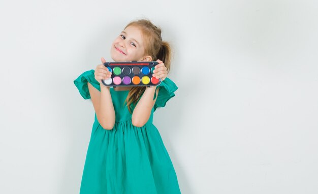 Little girl in green dress holding watercolor paints with brush and looking cheerful