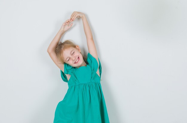 Маленькая девочка в зеленом платье протягивает руки с закрытыми глазами и выглядит весело
