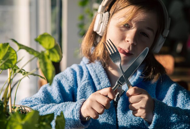A little girl in headphones and holds knife with a fork in her hands