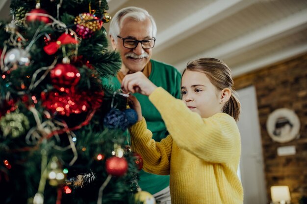 Маленькая девочка и ее дедушка украшают елку дома