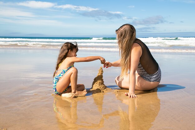 Маленькая девочка и ее мама строят замок из песка на пляже, сидя на мокром песке, наслаждаясь отпуском на море